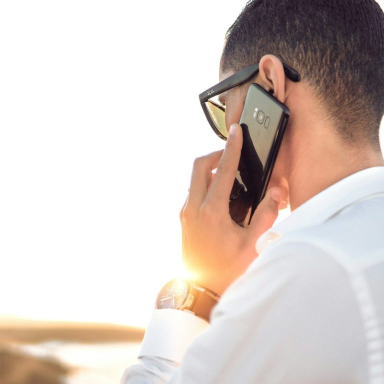 Mann mit Sonnenbrille telefoniert und schaut in die Ferne, Sonnenuntergang im Hintergrund.