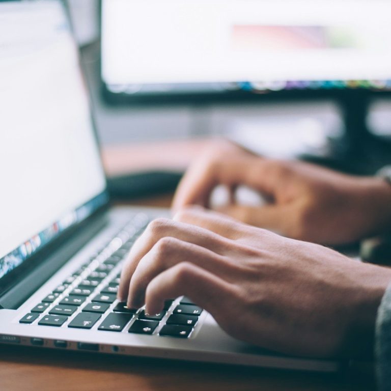 Hände tippen auf einer Laptop-Tastatur, im Hintergrund ein Computerbildschirm.