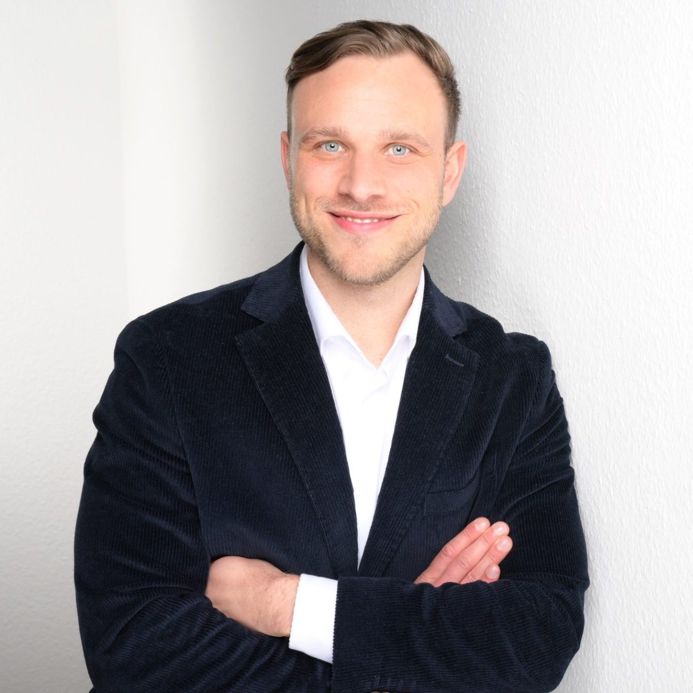 Mann mit lächelndem Gesicht, in schwarzem Sakko und weißem Hemd, an einer Wand stehend.